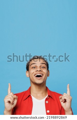 Young smiling fun man of African American ethnicity 20s wears red shirt point index finger overhead indicate on workspace area copy space mock up isolated on plain pastel light blue cyan background