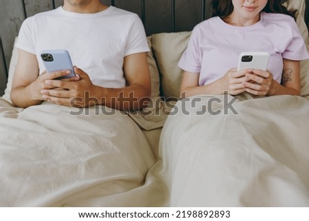 Cropped young couple two family man woman she he wearing t-shirt pajama hold use mobile cell phone peeps sitting in bed rest spend time together in bedroom home in own room hotel. Real estate concept