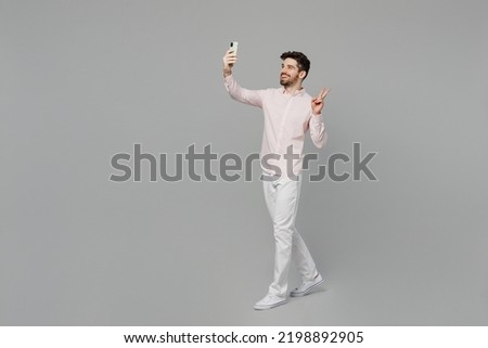 Full body young happy caucasian man 20s he wearing basic white shirt doing selfie shot on mobile cell phone post photo on social network show v-sign isolated on plain grey background studio portrait