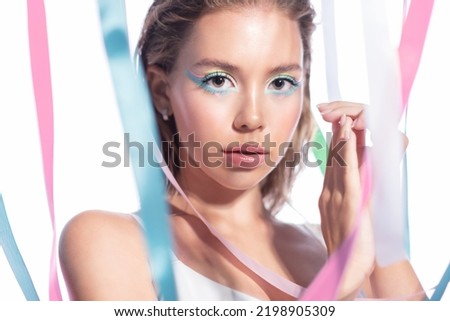 Beautiful blonde model with wet hair with makeup in blue tones on the background of fluttering satin ribbons in soft pastel colors looks into the camera on a white background