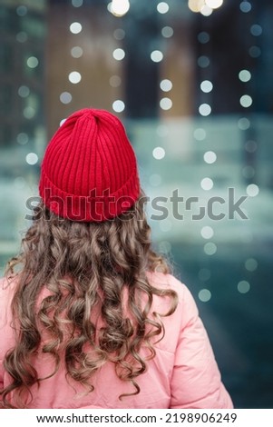 Winter mood: girl or woman  with long curly light hair in red hat stands with her back to the camera, looking at the blurred window, lights bokeh background, outdoors, vertical