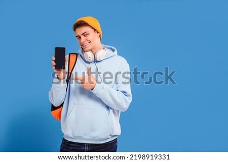 Photo of young fashionable student guy with cap and backpack smiling at camera, pointing on empty screen of mobile phone, isolated over blue studio background. A lot of copy space. 
 Royalty-Free Stock Photo #2198919331