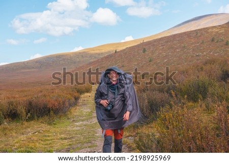 Alone traveler in raincoat with large backpack walks along hiking trail among fading autumn vegetations on high mountain plateau. Backpacker with photo camera in mountain trekking in rainy weather.