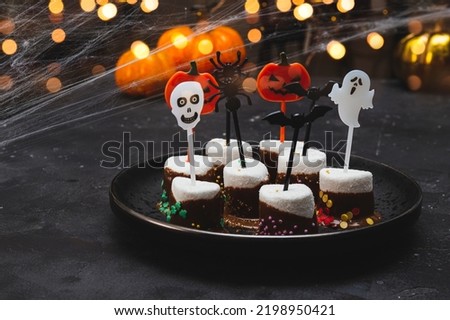  Sweet Halloween treat, marshmallows covered chocolate with sugar and Halloween decorations on a black plate, photo with soft focus, golden bokeh