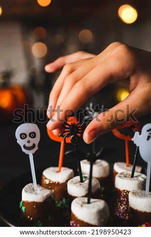 Hand is taking sweet Halloween treat, marshmallows covered chocolate with sugar and Halloween decorations on a black plate, vertical photo with soft focus, golden bokeh