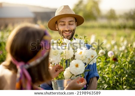 Portrait of cheerful man and woman stand as farmers with buckets full of freshly picked up dahlias while working at flower farm outdoors on sunset Royalty-Free Stock Photo #2198963589