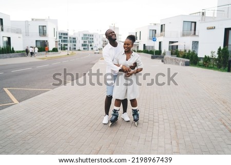 Full body of laughing Dark Skinned boyfriend having fun while teaching happy girlfriend in rollerblades skate on city street during date