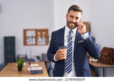 Young hispanic man business worker talking on smartphone drinking coffee at office