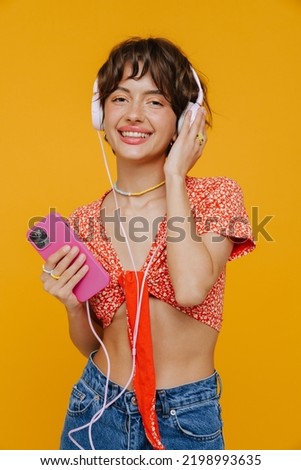 Young beautiful smiling short-haired stylish woman in headphones with phone looking at camera while standing with her side over isolated yellow background
