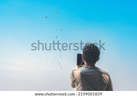 Male parents take video clips and photo with smart phone and watch with worry and concern during parachute training from airplane for army cadet with blurred image of parachute and sky in background.