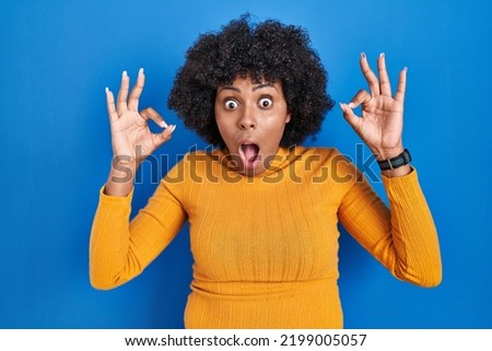 Black woman with curly hair standing over blue background looking surprised and shocked doing ok approval symbol with fingers. crazy expression 