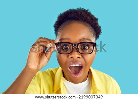 Wow, just look at that. Headshot of beautiful nerdy young African American woman in yellow shirt and glasses isolated on blue background staring at something with happy, surprised face expression