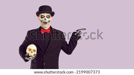 Man in Halloween costume with surprised, scared and shocked expression is pointing at advertising space. Man in suit and Halloween make-up holds skull and points to copy space on lilac background.