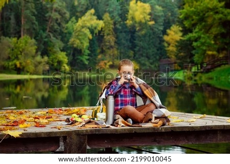 the boy is sitting wrapped in a blanket on a wooden pier next to the lake and drinking hot tea from a thermos. the concept of a picnic in autumn, going out into nature. Royalty-Free Stock Photo #2199010045