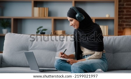 Islamic Arabian student girl Muslim woman wearing black hijab busy lady in headphones with laptop watching webinar lesson online class training write notes in notebook do homework e-learning at home