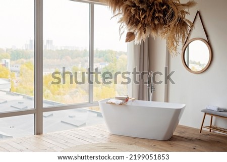 Comfortable bright bathroom with a boho-chic interior design, a free-standing white bath against the background of a panoramic window. hanging decor from dry grass. Soft selective focus. Royalty-Free Stock Photo #2199051853
