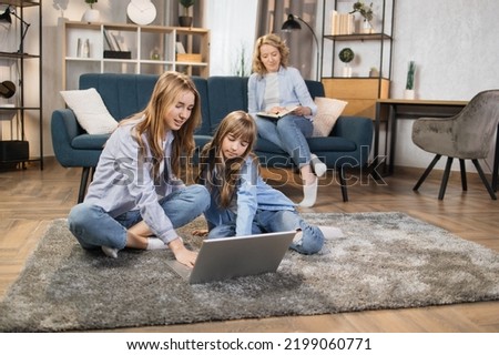 Cute blond girls sisters sitting on warm floor play at home together using laptop, while happy mother relax on sofa on background.