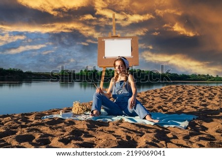 A woman during the open air on the sandy bank of the river with an easel. An artist with paints near an empty mock up sheet in nature.