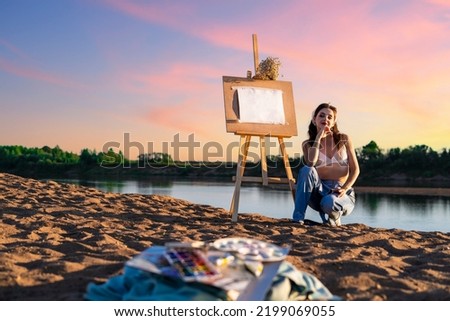 A woman during the open air on the sandy bank of the river with an easel. An artist with paints near an empty mock up sheet in nature.