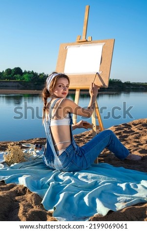 A woman during the open-air on the sandy bank of the river with an easel. An artist with paints near an empty mock up sheet in nature.