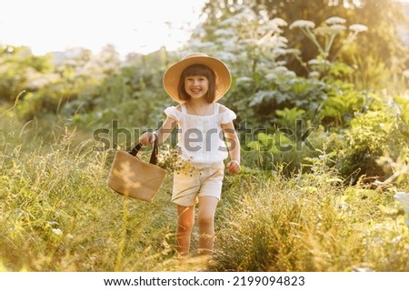 Portrait of smiling little girl walks in the rays of a sunset in a flowering meadow, enjoying the summer, warmth, flowers, freedom. Child with straw hat and bag is having fun outdoors. Royalty-Free Stock Photo #2199094823