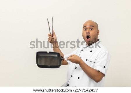Young asian male chef in uniform cooking with iron grill pan and tongs kitchen utensil. Cooking man Occupation chef People in kitchen restaurant and hotel.