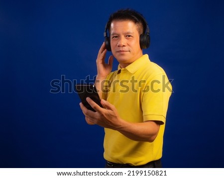 Portrait of smart middle-aged asian man in casual yellow T-shirt listening music with wireless headphone and smartphone isolated on blue background in studio, looking happiness and attractive
