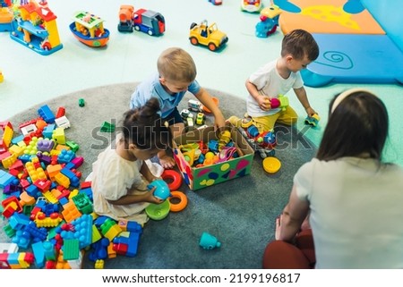 Toddlers and their nursery teacher playing with plastic building blocks and colorful car toys in a nursery school playroom. Early brain and skills development. High quality photo