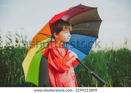 Portrait of a smiling school boy with rainbow umbrella in the park. Kid holds colourful umbrella on his shoulder. Cheerful child in a red raincoat holding multicolor umbrella outdoors. Royalty-Free Stock Photo #2199215899