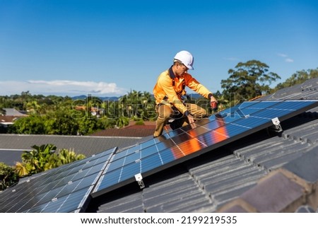 Solar panel technician with drill installing solar panels on house roof on a sunny day. Royalty-Free Stock Photo #2199219535