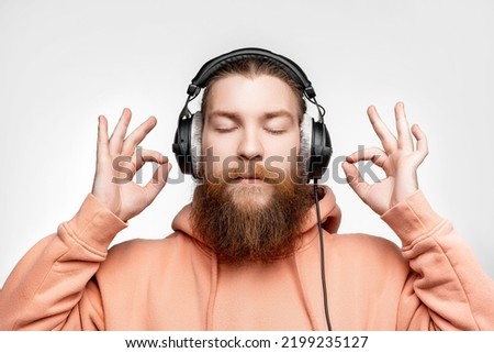 Scandinavian handsome man shows ok gesture keep calm and listens to music in professional headphones isolated on gray background. Happy guy with ginger hairstyle and beard. Modern digital technologies