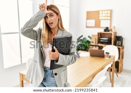 Blonde business woman at the office making fun of people with fingers on forehead doing loser gesture mocking and insulting. 
