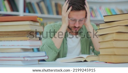 A tired student sits at desk full of books. Young man preparing for an exam in the college library. Self education concept.