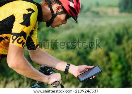 Happy male cyclist sending a text message on his mobile phone. Image of a cyclist on a mountain bike using a smartphone navigator exploring a map and searching for GPS coordinates while cycling