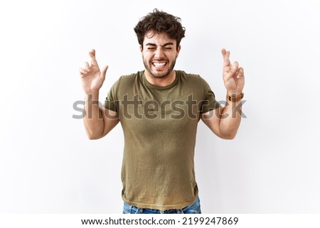 Hispanic man standing over isolated white background gesturing finger crossed smiling with hope and eyes closed. luck and superstitious concept. 