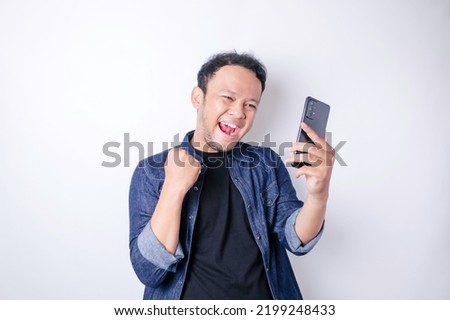 A young Asian man with a happy successful expression wearing navy blue shirt and holding smartphone isolated by white background