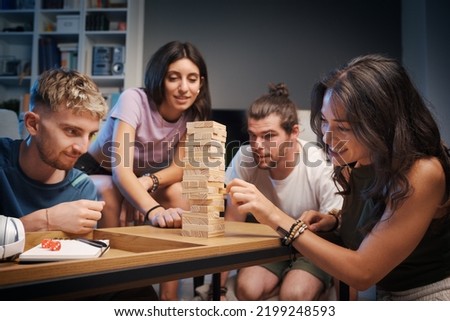 Young happy people playing Jenga at home, a girl is removing a wooden block from the tower and the others are watching Royalty-Free Stock Photo #2199248593