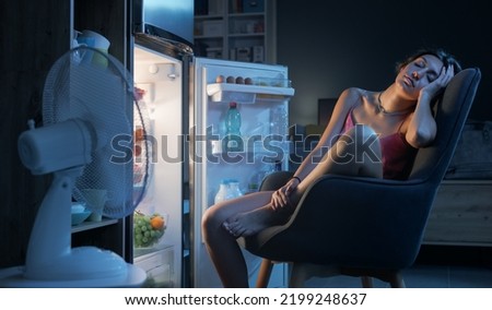 Exhausted woman suffering from the heat during the summer heatwave, she is sitting in front of the open fridge and cooling herself Royalty-Free Stock Photo #2199248637
