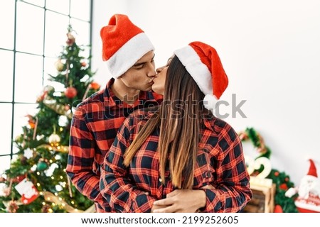 Young latin couple kissing and hugging standing by christmas tree at home.