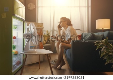 Angry your woman sitting in front of the open fridge and talking on the phone, her air-conditioning system is broken Royalty-Free Stock Photo #2199253021