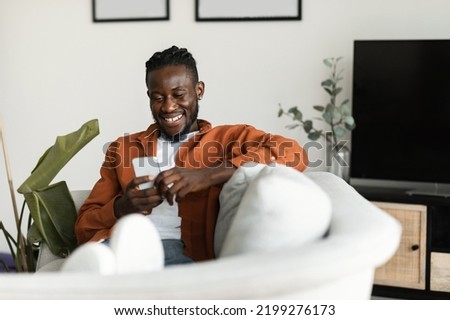 Cool gadget and app. Excited black man holding mobile phone and typing sms message, resting on sofa at home, free space. Man browsing internet and surfing web Royalty-Free Stock Photo #2199276173