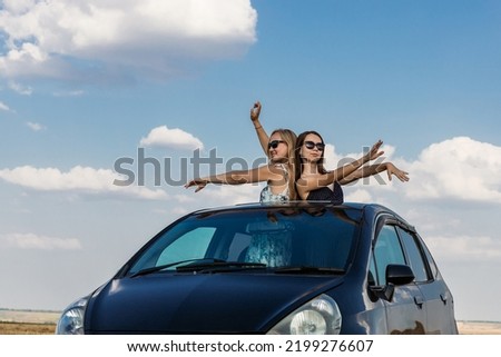 Beautiful young women in sunglasses dance against the background of a blue sky with clouds leaning out of the car hatch. The concept of traveling by private car Royalty-Free Stock Photo #2199276607