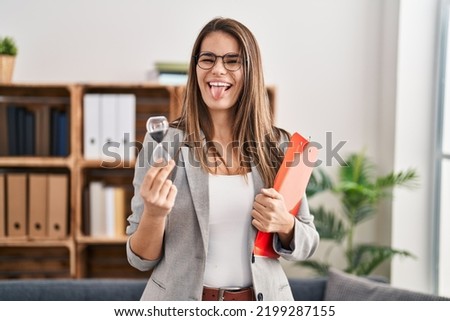 Beautiful woman working at therapy office holding sand clock sticking tongue out happy with funny expression. 