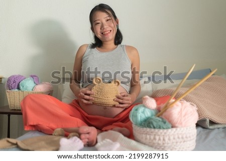 lifestyle home portrait of young happy and beautiful Asian Chinese woman knitting little bonnet for the new baby relaxed in her bedroom in maternity and handicraft concept Royalty-Free Stock Photo #2199287915