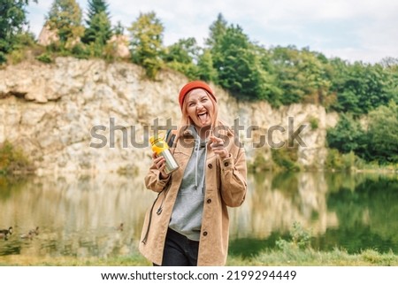 Smiling, positive blonde woman in stylish clothes holding a thermos and looking at the camera, posing showing a peace sign on the cold forest lake in Poland. 