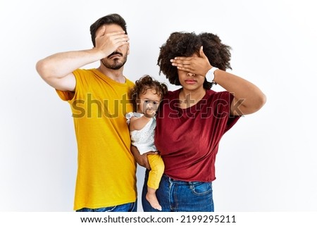 Interracial young family of black mother and hispanic father with daughter covering eyes with hand, looking serious and sad. sightless, hiding and rejection concept 