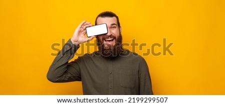 Playful bearded man covers one eye with phone screen and smiles at the camera. Studio portrait over yellow background.