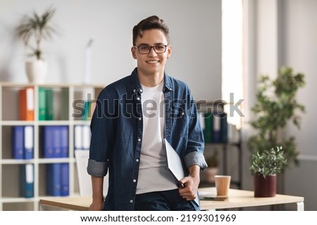 Portrait Of Handsome Young Male Entrepreneur Holding Laptop And Posing In Office Interior, Confident Millennial Man In Casual Clothes Standing Near Desk And Smiling At Camera, Copy Space Royalty-Free Stock Photo #2199301969