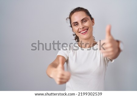Beautiful brunette woman standing over isolated background approving doing positive gesture with hand, thumbs up smiling and happy for success. winner gesture. 