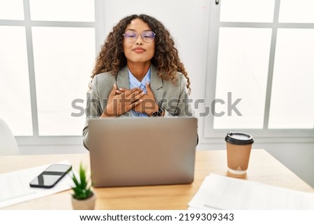 Beautiful hispanic business woman sitting on desk at office working with laptop smiling with hands on chest with closed eyes and grateful gesture on face. health concept. 